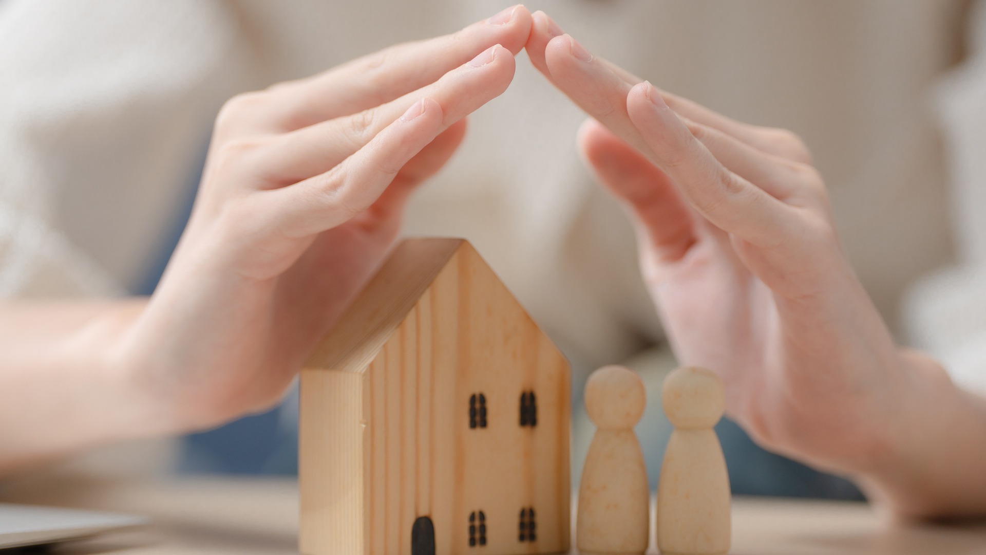 A pair of hands covering a wood plank in the shape of a house and two wood figures.