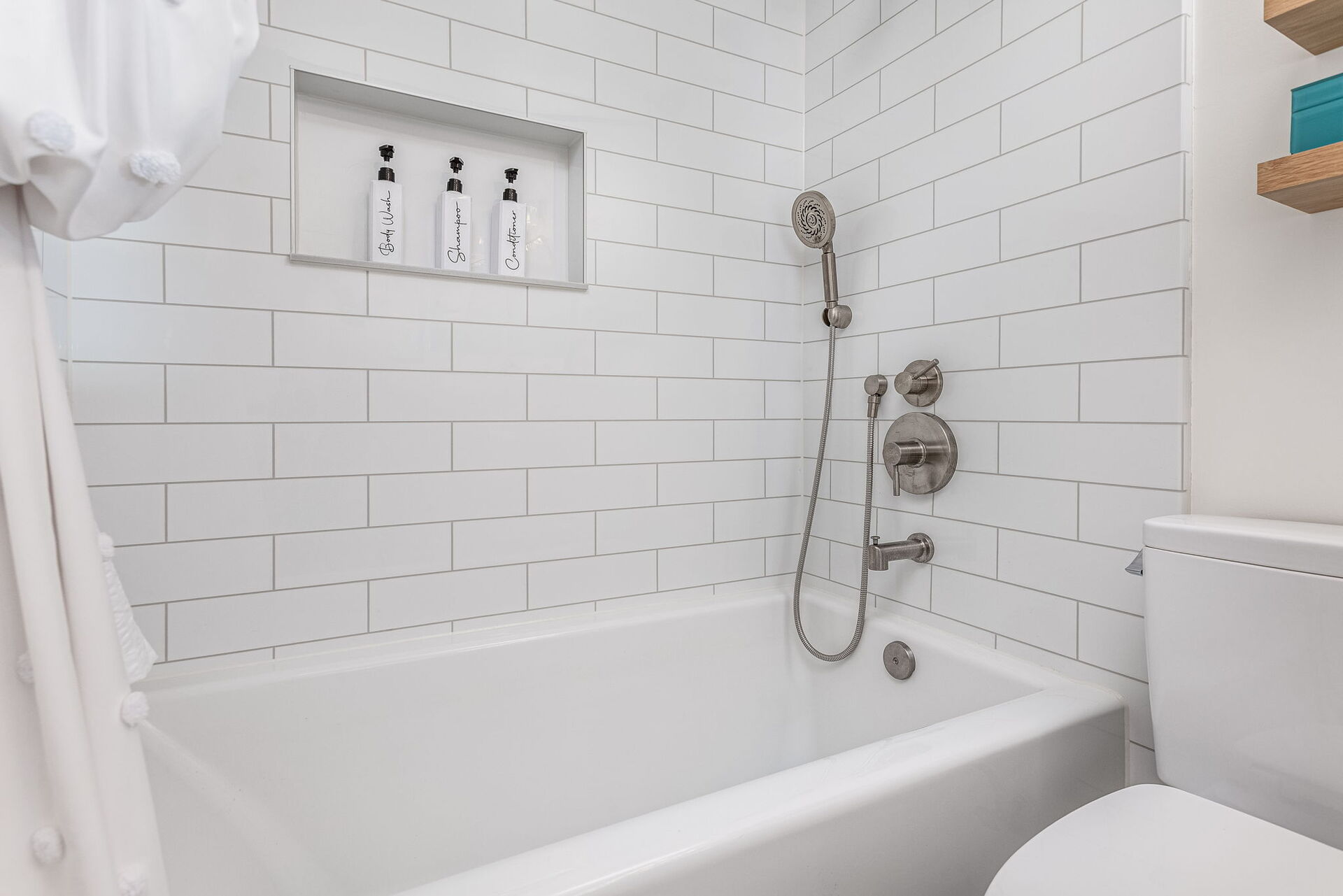 A remodeled shower-tub combo with tile-pattern walls, a handheld shower, and a storage niche with three shampoo bottles.