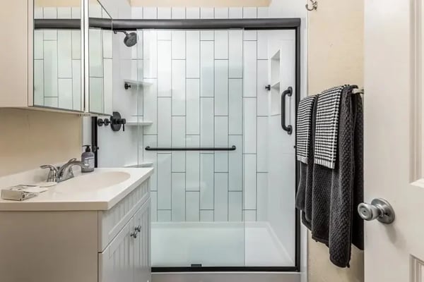 A freshly remodeled bathroom featuring a sleek walk-in shower. The shower includes a built-in corner shelving unit, a recessed niche for storage, and matte black hardware throughout – including a grab bar, fixtures, and a sliding glass door frame. The vanity area is simple and clean, with a white countertop and mirrored cabinet.