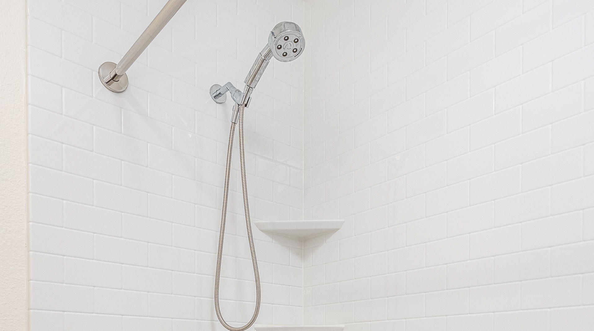 A remodeled shower with a handheld chrome shower head, a tile-like white surround, and two shelves.