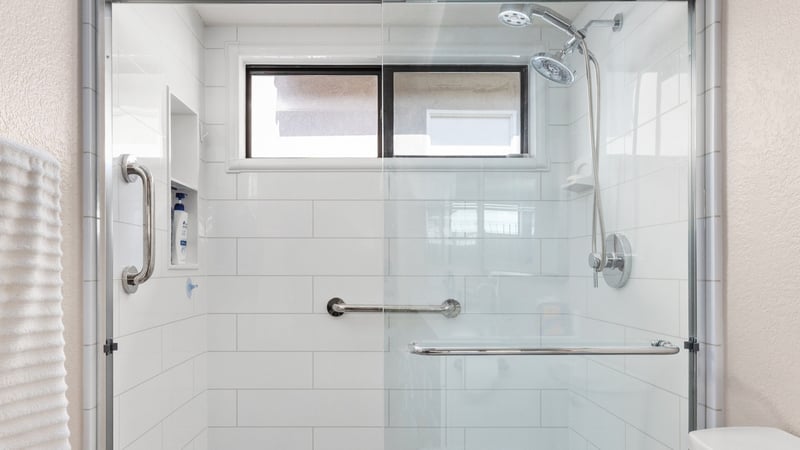 A shower remodel with two grab bars, a shower niche, clear glass, and a handheld-fixed shower head combo