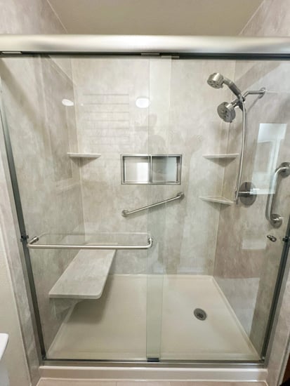 A remodeled walk-in shower with clear sliding glass doors, a handheld-fixed shower head combo, two grab bars, three corner shelves, a shower niche, and a shower bench