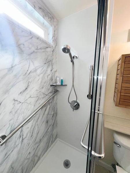 A walk-in shower with a handheld shower head, two grab bars, clear glass, and a Calcutta marble shower wall