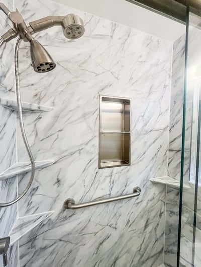 A shower with Calcutta marble walls, a fixed and hose shower head, a grab bar, four corner shelves, and one double shower niche.
