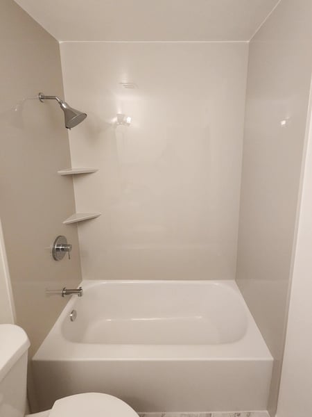 A tub and shower combo with plain walls, two corner shelves, and a fixed shower head.