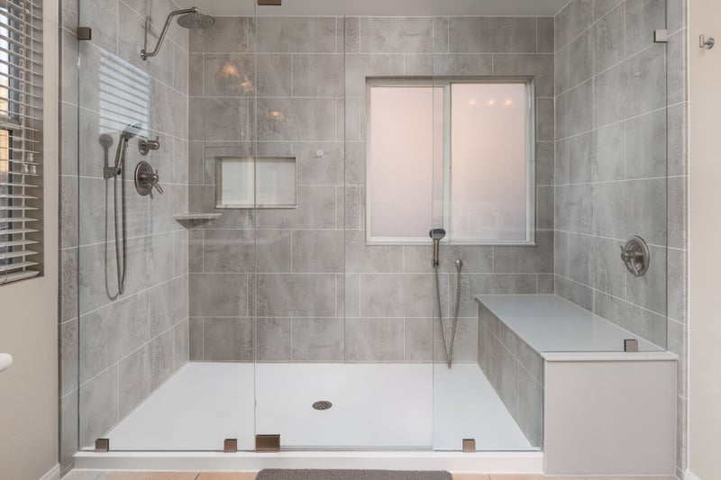 A new walk-in shower with clear glass doors, a custom shower seat, two handheld shower heads, one rain shower head, a corner shelf, and a storage niche
