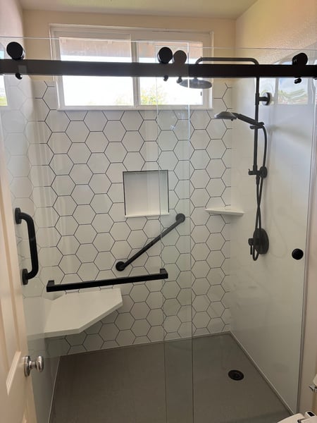 A walk-in shower with a hexagonal pattern accent wall, shower storage niche, two grab bars, and a sliding glass door