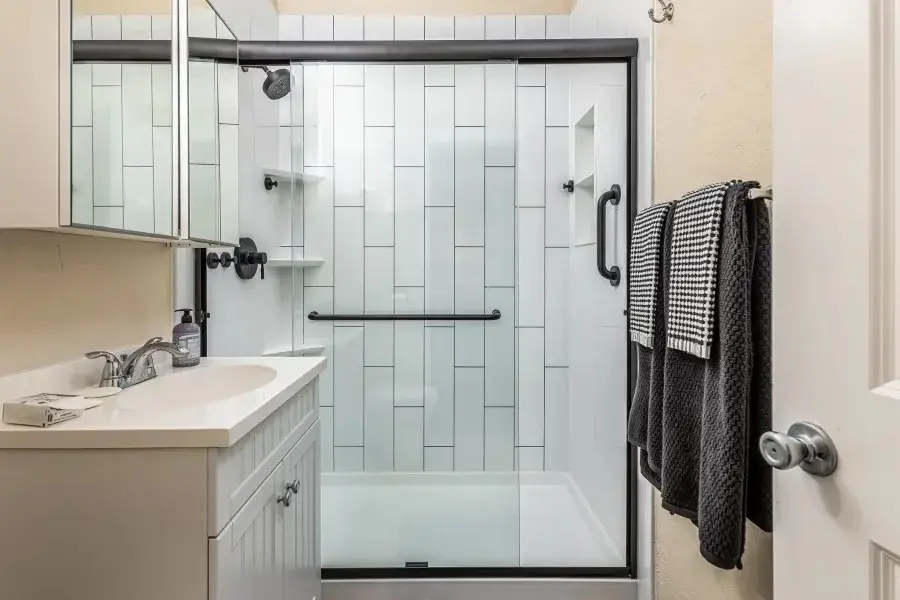 A freshly remodeled bathroom featuring a sleek walk-in shower. The shower includes a built-in corner shelving unit, a recessed niche for storage, and matte black hardware throughout – including a grab bar, fixtures, and a sliding glass door frame. The vanity area is simple and clean, with a white countertop and mirrored cabinet. 