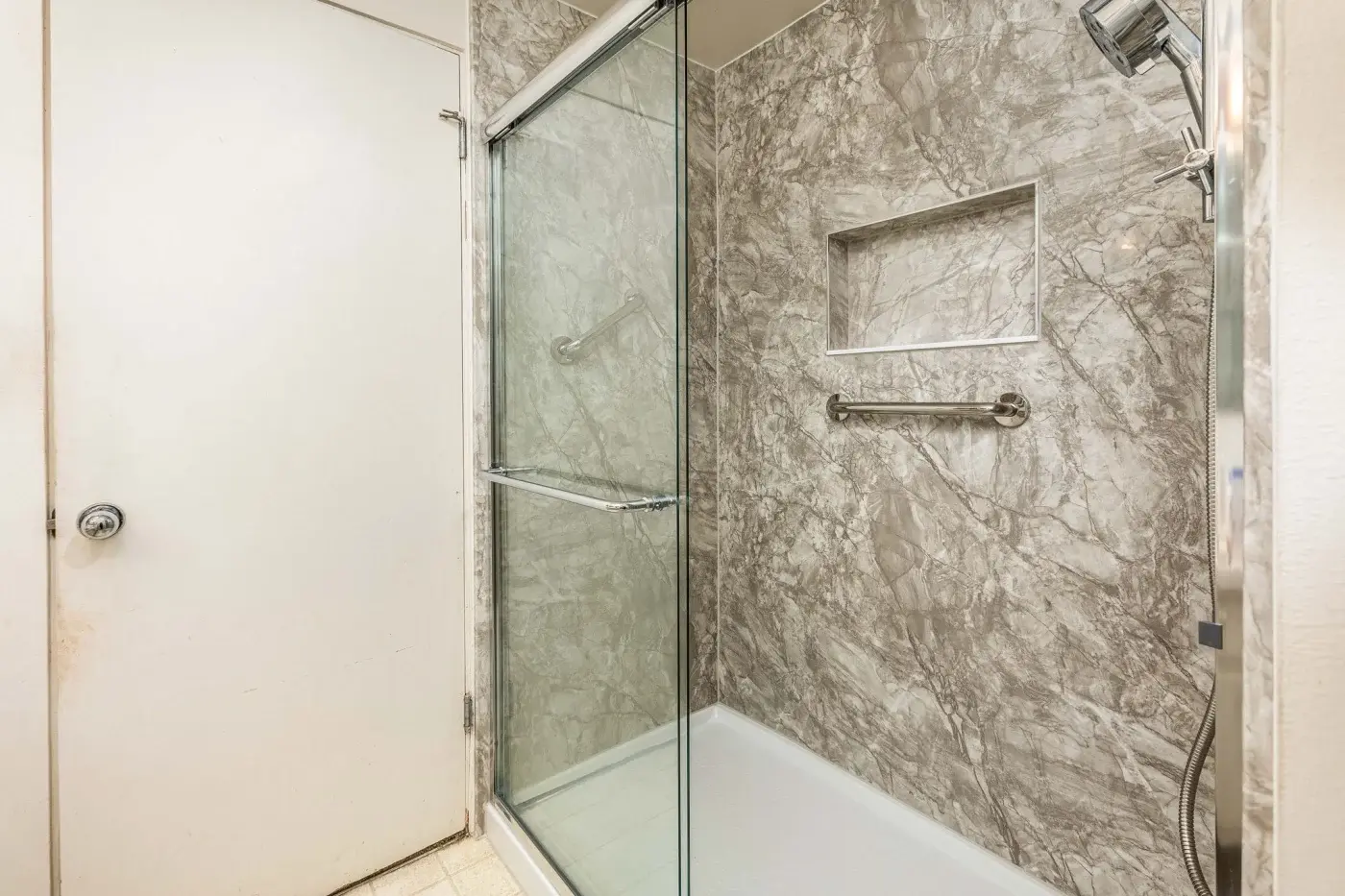 A newly remodeled walk‑in shower by Shugarman’s Bath, featuring elegant, marble‑look wall panels in shades of gray and white. The shower is enclosed with sliding glass doors framed in polished chrome, matching the chrome fixtures throughout. A built‑in recessed niche provides convenient storage for bath products, while a horizontal grab bar adds safety and accessibility. The showerhead is mounted on the right wall, with a handheld attachment for added versatility. The clean, grout‑free wall surface offers a modern, low‑maintenance upgrade that blends both style and function.